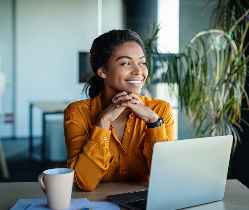 A woman in a yellow blouse sits at her laptop researching 2024 cybersecurity predicitons with a cup of coffee looking out the window.